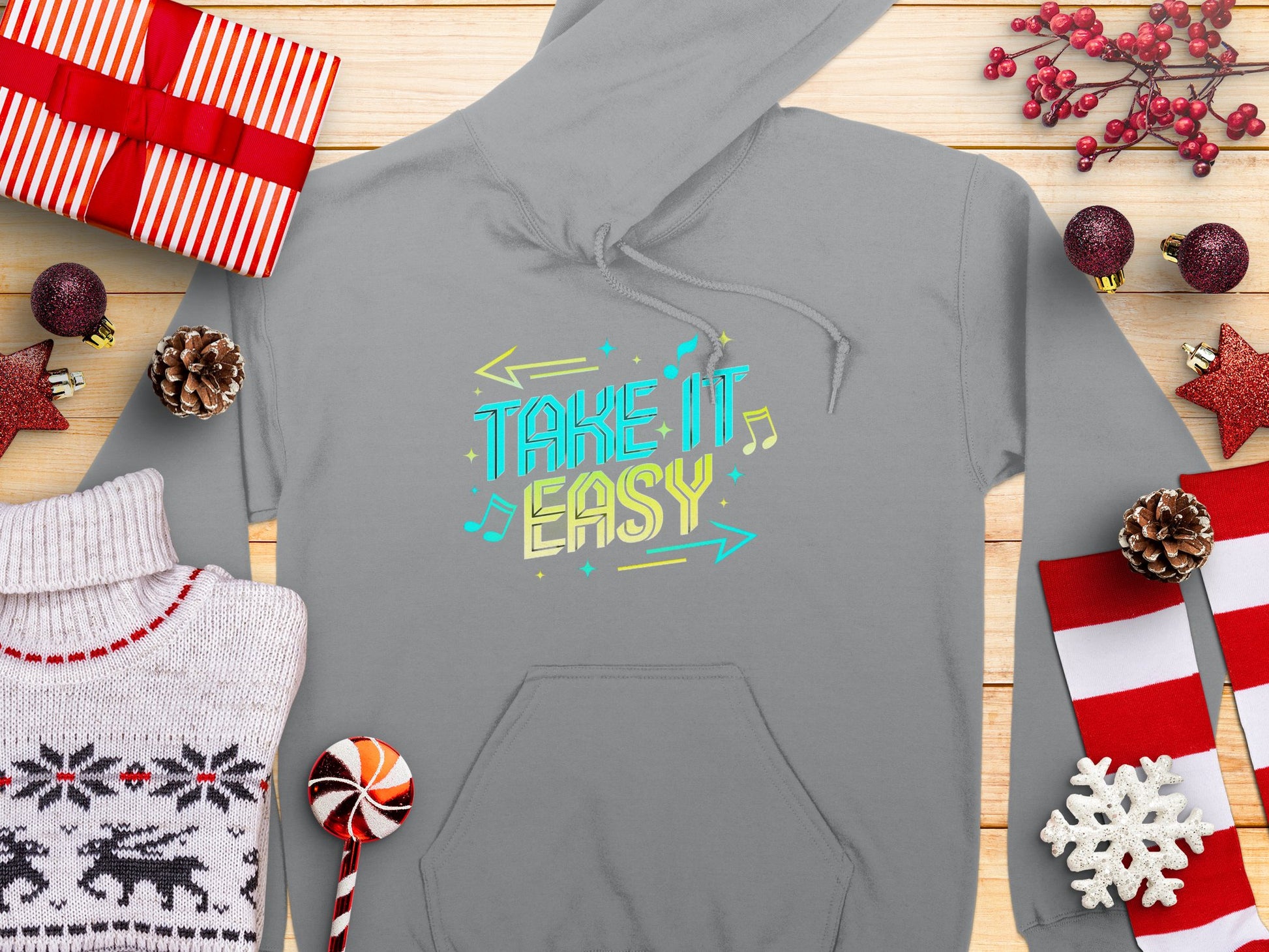 A gray hoodie from Garment Graphics with Take It Easy in colorful letters lays on a wooden surface, creating a cozy fashion statement. Its surrounded by wrapped gifts, red and white striped ribbons, pine cones, Christmas ornaments, a candy cane, and a knitted sweater.