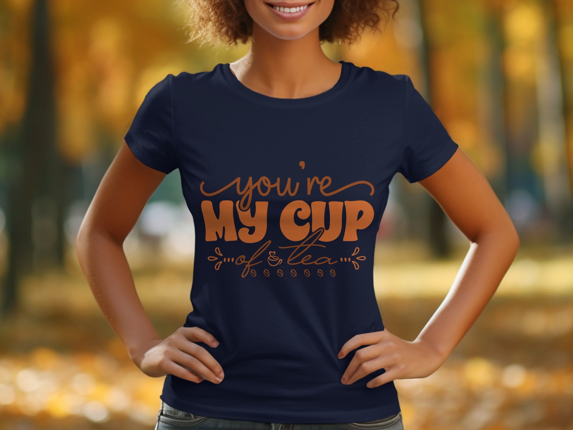 A person stands outdoors wearing a navy 100% cotton T-shirt by Garment Graphics, featuring Youre My Cup of Tea in orange cursive. The background is a blurred autumnal scene with trees and fallen leaves.