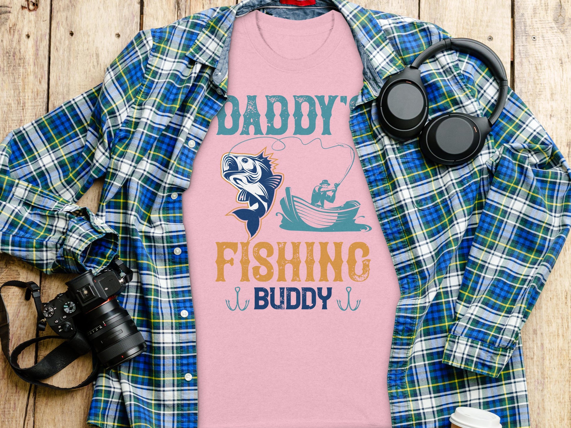 Garment Graphics Daddys Fishing Buddy graphic print T-shirt, featuring a fish and angler in a boat design, is displayed on a wooded surface over a checked shirt. Ideal for fishing enthusiasts, it is accompanied by headphones, a camera, and a white to-go cup.