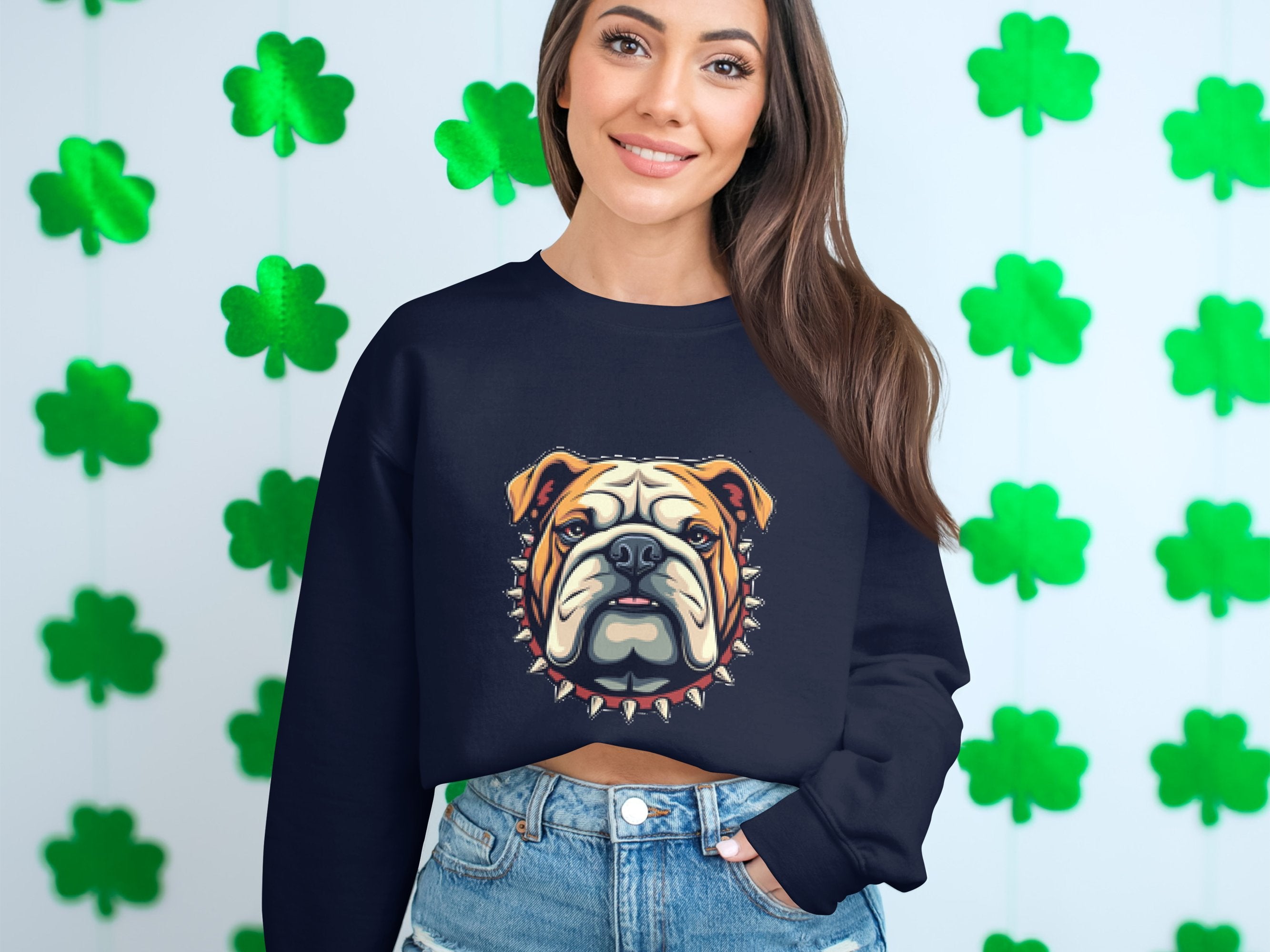 A person poses in front of a wall full of green shamrock cutouts, wearing denim shorts and a black Garment Graphics sweatshirt with a bulldog design.