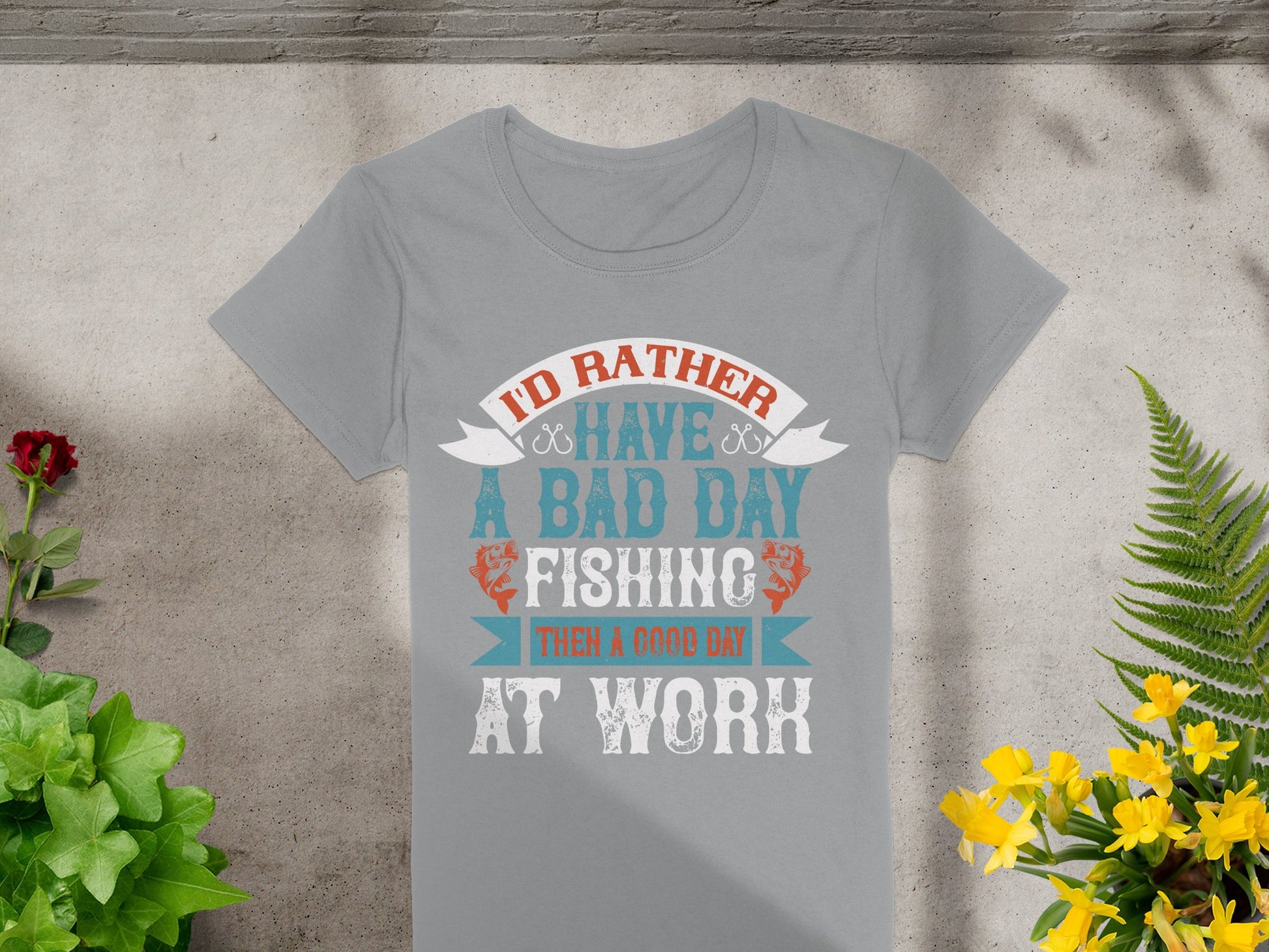 A Garment Graphics gray fishing T-shirt is displayed on a textured surface, featuring the playful text, Id rather have a bad day fishing than a good day at work. Designed for fishing enthusiasts in womens sizing, its encircled by green plants and yellow flowers.