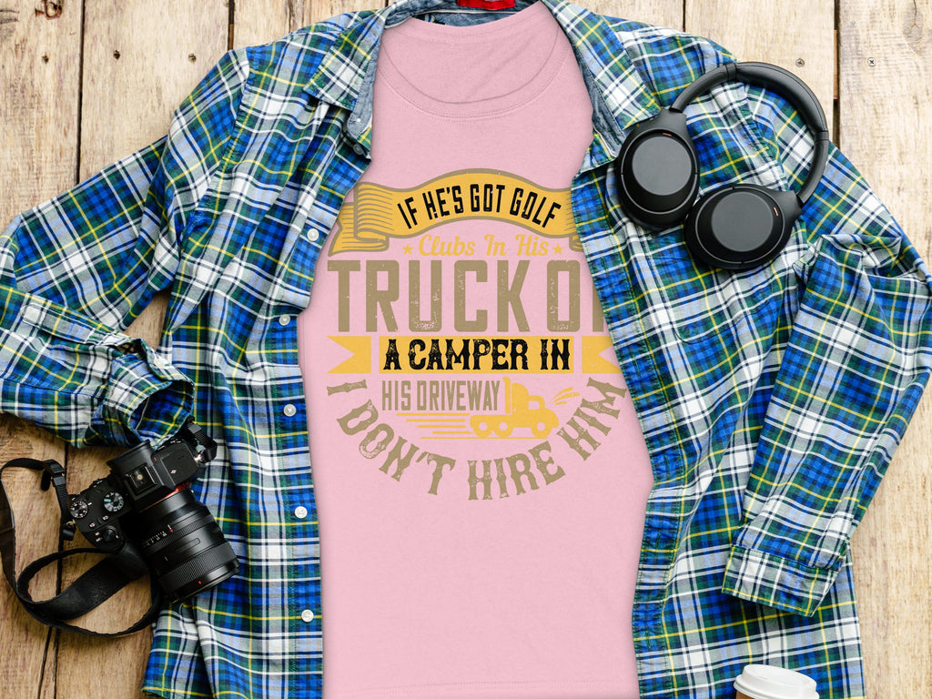 A quirky Garment Graphics pink womens t-shirt with the funny golf-themed text: If hes got golf clubs in his truck, a camper in his driveway, dont hire him rests on a wooden surface alongside a blue and green plaid shirt, headphones, and a camera.