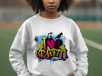 On a sports field, someone wears an artistic Garment Graphics sweatshirt featuring I Love Graffiti in colorful graffiti-style text, highlighting a pink heart with splashes of blue and yellow paint in an urban design.