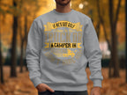 A person wearing a light gray Garment Graphics sweatshirt with gold text that reads, If hes got golf clubs in his truck or a camper in his driveway, don’t hire him, stands against tall trees with autumn foliage, embracing the outdoors.