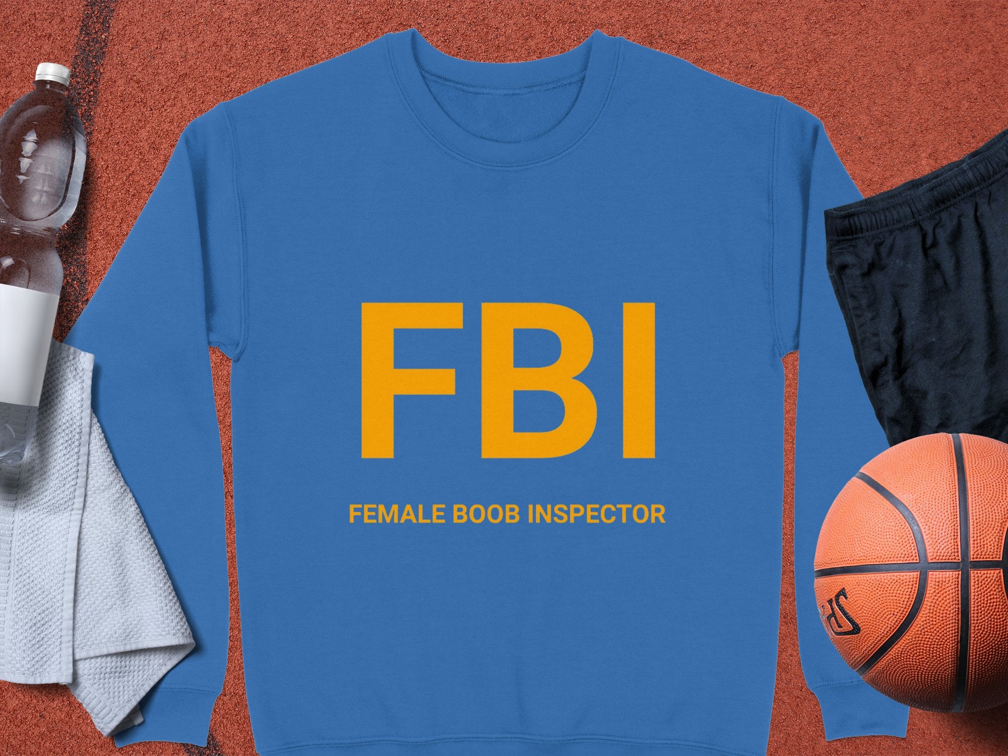 Garment Graphics offers a blue unisex sweatshirt featuring bold yellow FBI (Female Boob Inspector) letters. Set on a red surface, it complements a basketball, black shorts, towel, and water bottle.