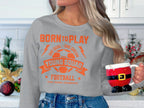 A person with long hair holds a Santa-themed mug, dressed in a Garment Graphics gray football sweatshirt with orange BORN TO PLAY YOUNG SQUAD FOOTBALL text. Festive decor enhances the mood, and the unisex sizing makes it perfect for anyone to enjoy the holiday spirit.
