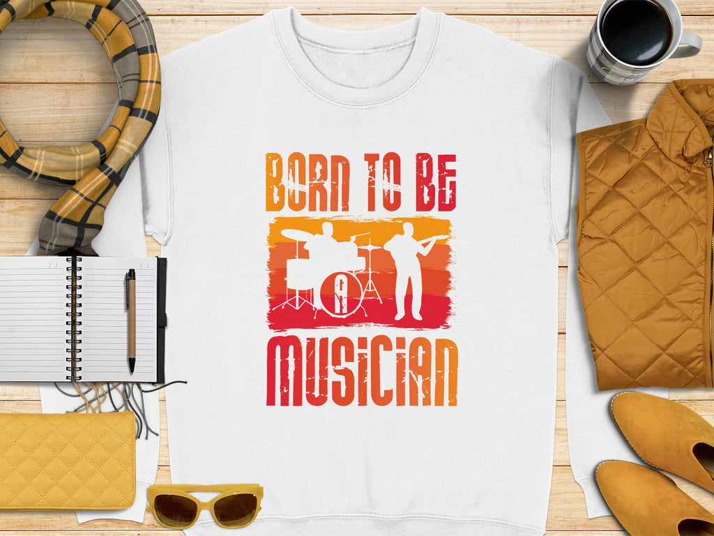 A white T-shirt by Garment Graphics has orange and red graphics with drum and guitar silhouettes and the phrase Born to Be Musician, ideal for music lovers. Pair it with a striped scarf, notebook, pen, yellow sunglasses, coffee cup, quilted jacket, and yellow wallet. Unisex sizing available.
