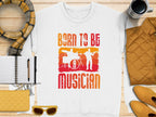 A white T-shirt by Garment Graphics has orange and red graphics with drum and guitar silhouettes and the phrase Born to Be Musician, ideal for music lovers. Pair it with a striped scarf, notebook, pen, yellow sunglasses, coffee cup, quilted jacket, and yellow wallet. Unisex sizing available.