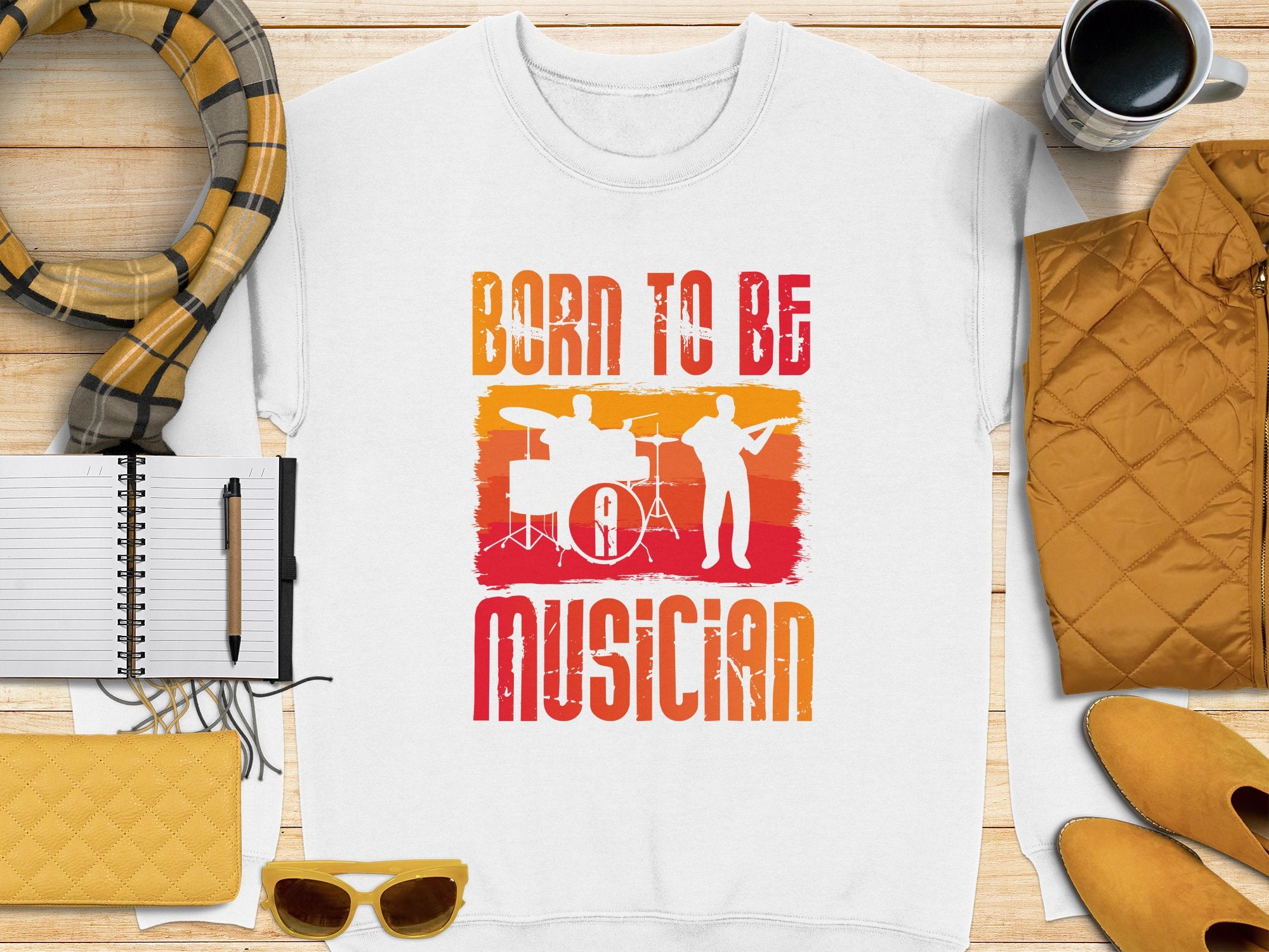 A white T-shirt by Garment Graphics has orange and red graphics with drum and guitar silhouettes and the phrase Born to Be Musician, ideal for music lovers. Pair it with a striped scarf, notebook, pen, yellow sunglasses, coffee cup, quilted jacket, and yellow wallet. Unisex sizing available.