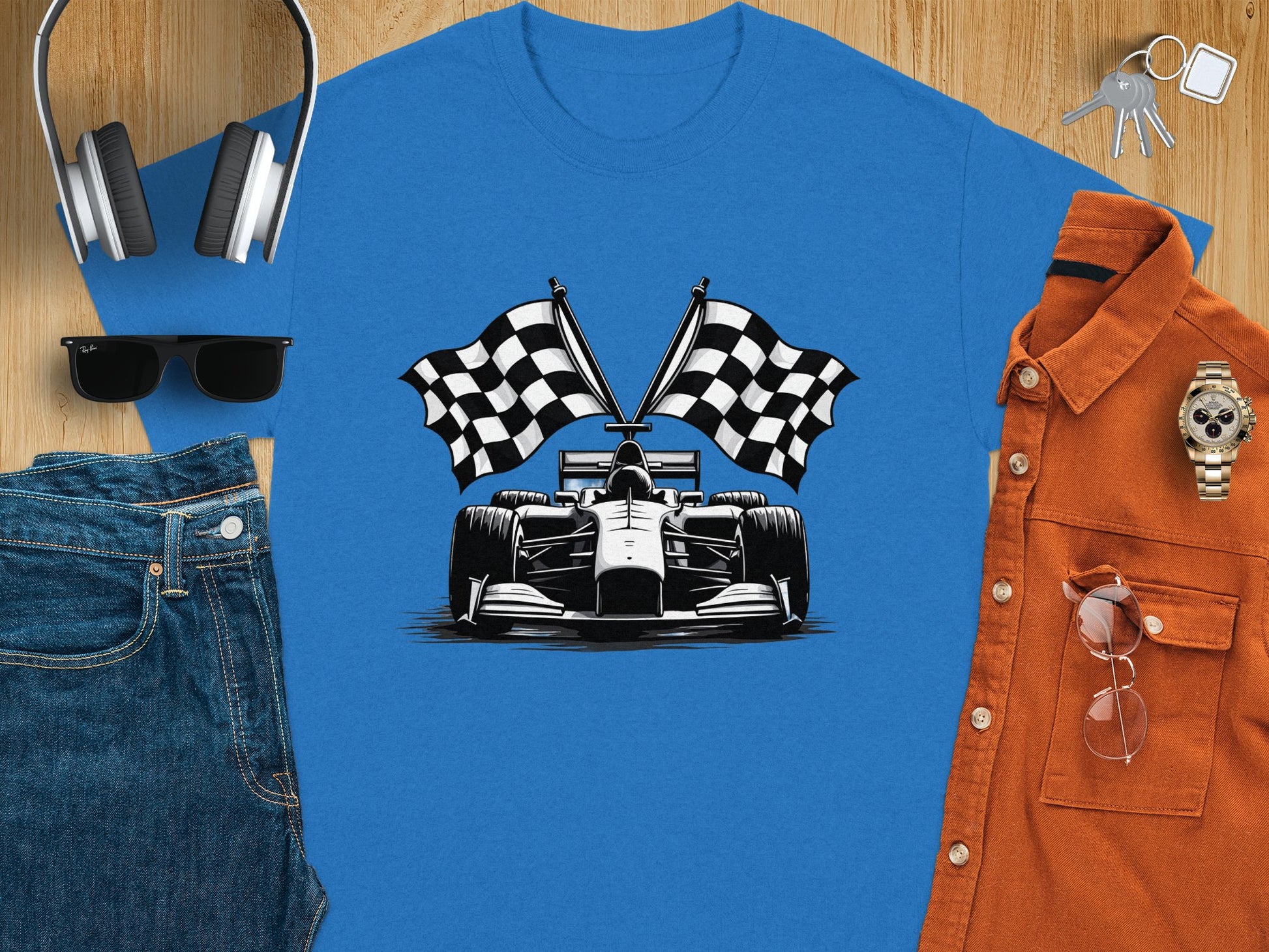 Flat lay showcasing Garment Graphics blue graphic T-shirt with race car and checkered flags, paired with jeans, sunglasses, headphones, keys, stylish watch, and an orange shirt on a wooden surface for a complete Formula One-inspired ensemble.