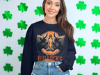 A woman with long brown hair smiles in a striking Garment Graphics navy blue sweatshirt featuring a BULLOCKS bullocks graphic. She pairs it with blue jeans against a backdrop of green shamrocks.