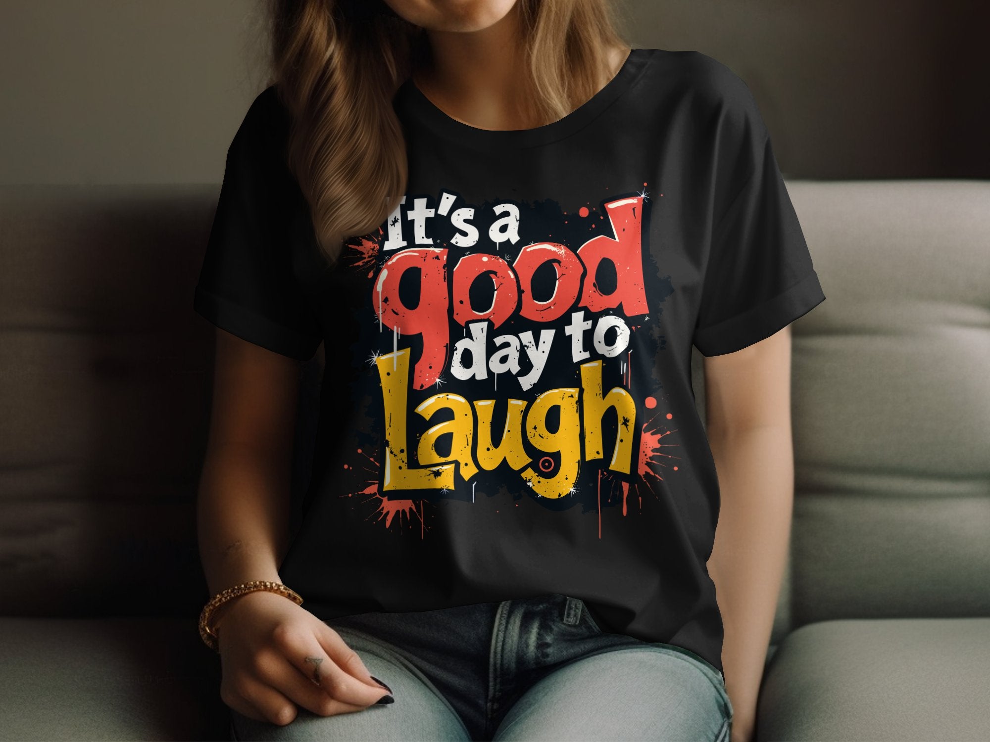 Someone is wearing a semi-fitted black t-shirt by Garment Graphics, showcasing a vibrant Its a good day to Laugh graphic. They sit on a gray couch with their hand gently resting on their lap, radiating positivity.