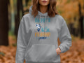 Wearing a Garment Graphics gray graphic hoodie with Daddys Fishing Buddy featuring a fish and boat, someone stands before a blurred forest scene with autumn foliage, ideal for outdoor adventures.