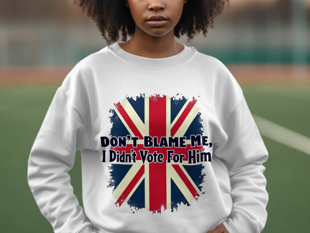 A person wears a Garment Graphics white unisex sweatshirt featuring a British flag and Dont Blame Me, I Didnt Vote For Him text, with a tennis court in the blurred background.