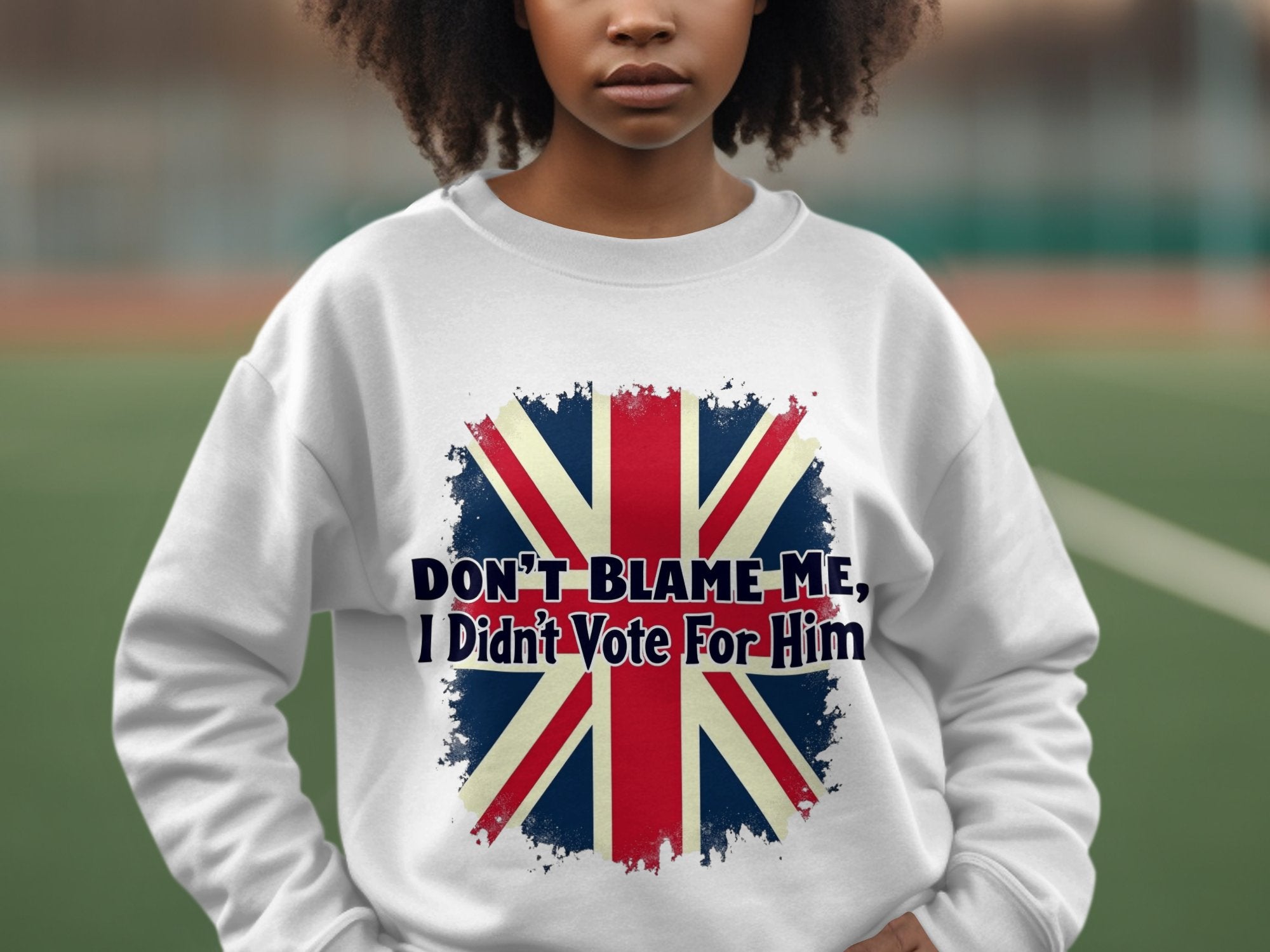 A person wears a Garment Graphics white unisex sweatshirt featuring a British flag and Dont Blame Me, I Didnt Vote For Him text, with a tennis court in the blurred background.