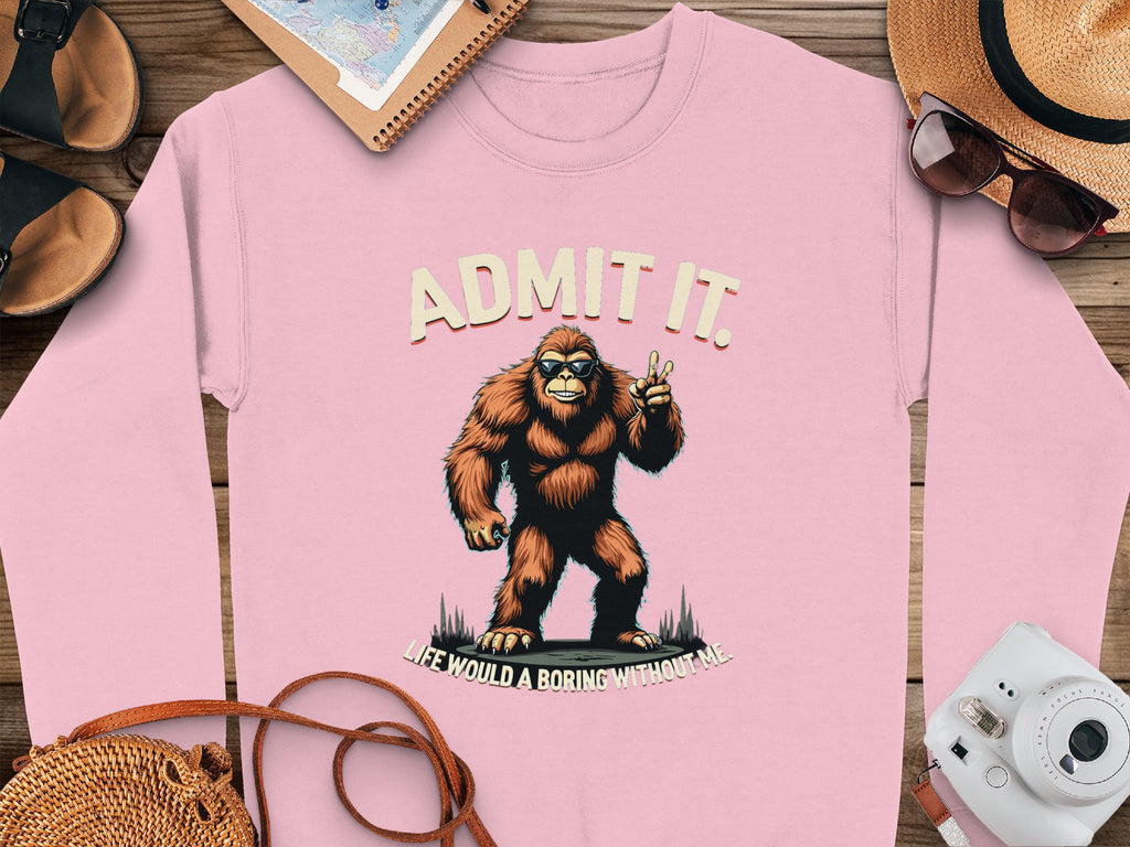 This pink Garment Graphics sweatshirt showcases a playful Bigfoot flashing a peace sign with the words Admit It above and Life Would Be Boring Without Me below. Unisex sizing makes it ideal for casual wear, paired with essentials like a camera, hat, sunglasses, notebook, and sandals.