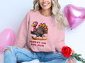 Someone is wearing Garment Graphics unisex pink sweatshirt featuring a humorous cartoon turkey with the text Turkey on the Run. Theyre sitting cross-legged, holding a red rose in front of pink heart-shaped balloons.