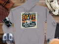 A Garment Graphics gray graphic hoodie crafted from medium-heavy fabric showcases a vibrant design of a person with blue, orange, and green text: Just Dave Doing Dave Things. Its artistically displayed on a wooden surface among a camera, binoculars, and other items.