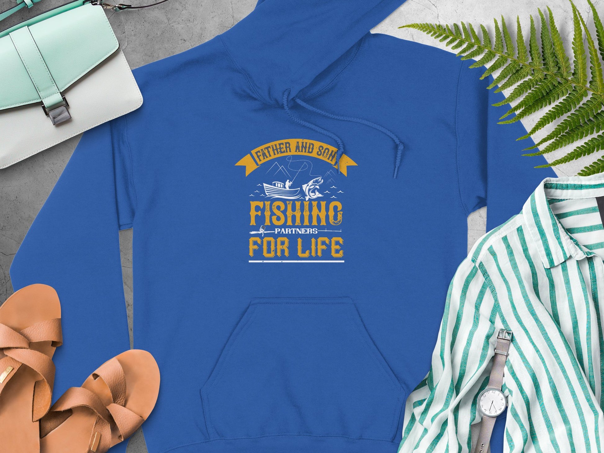 Garment Graphics blue hoodie, with Father and Son Fishing Partners for Life and boat/fish illustrations, enhances outfits alongside a green handbag, tan sandals, striped shirt, watch, and green fern leaves on a gray surface—a perfect pick for fishing lovers.