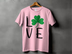 A pink T-shirt by Garment Graphics hangs on a wooden hanger, showcasing Irish spirit with L💚VE, where a green shamrock replaces the O. This charming St. Patricks Day shirt is set against a gray wall.