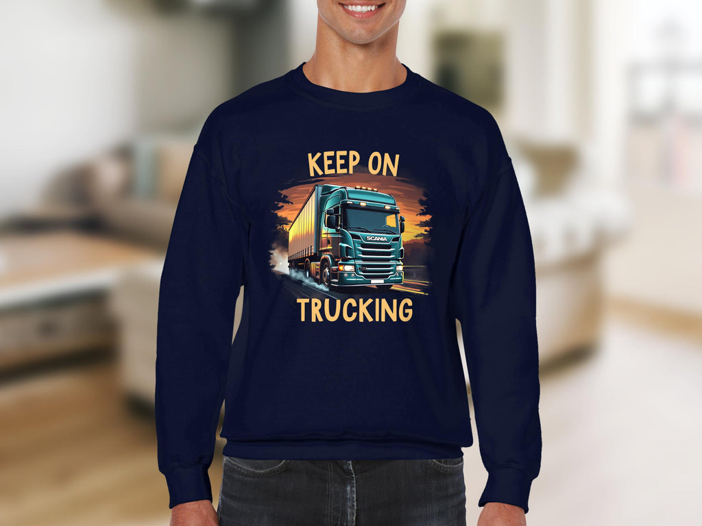A person sports a Garment Graphics navy sweatshirt featuring a truck illustration and the phrase Keep On Trucking, ideal for truck fans, with a blurred room interior in the background.