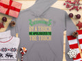 A humorous Garment Graphics hoodie lies on a wooden surface adorned with holiday gifts, pine cones, and candy. It displays the funny quote: If an Englishman gets run over by a truck, he apologizes to the truck in green, yellow, and white—perfectly capturing English etiquette.