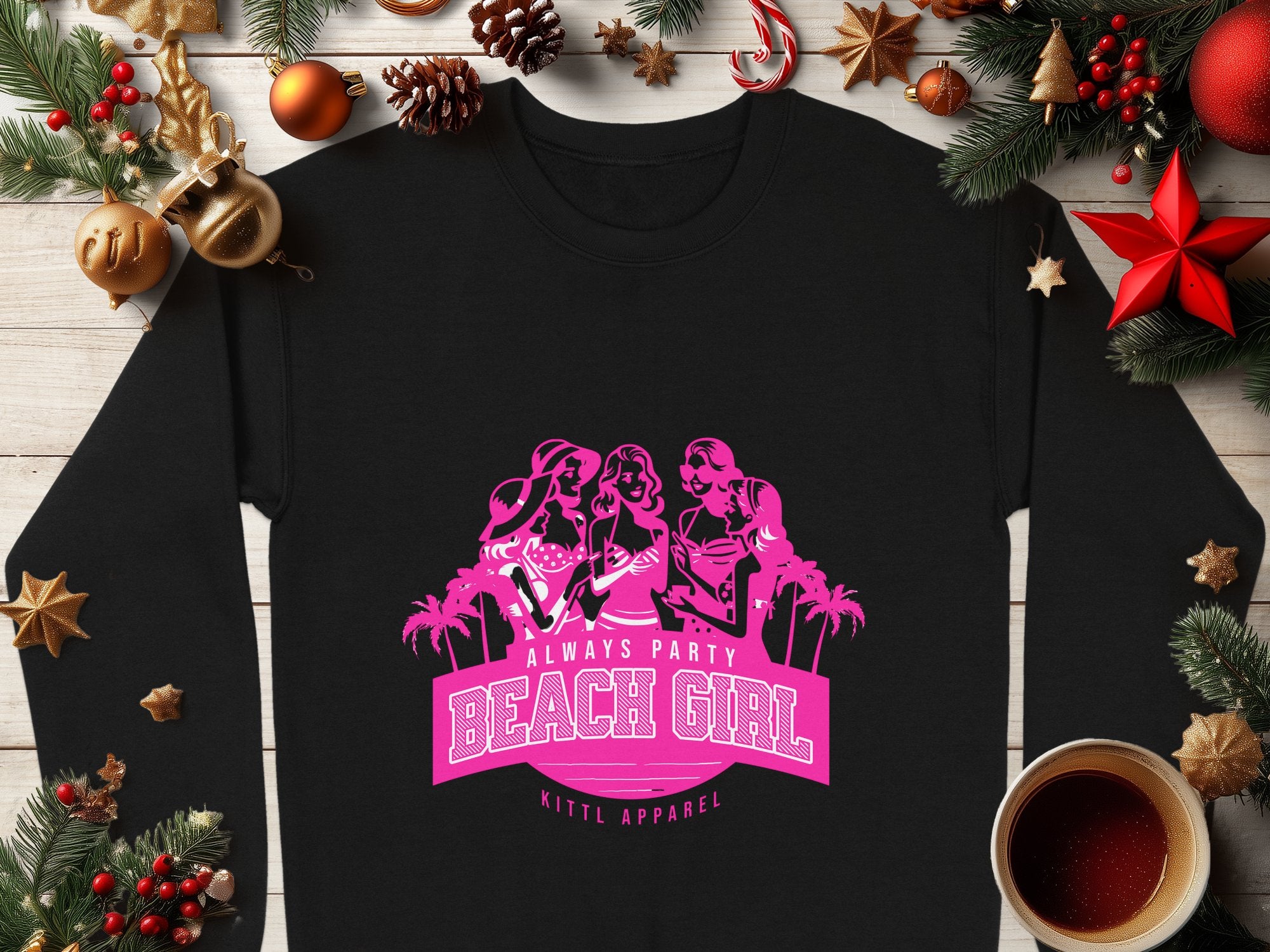 A black sweatshirt from Garment Graphics is on a wooden surface with Christmas decorations. It features a pink graphic of three women, palm trees, and the phrase ALWAYS PARTY BEACH GIRL. A steaming cup of coffee sits nearby.