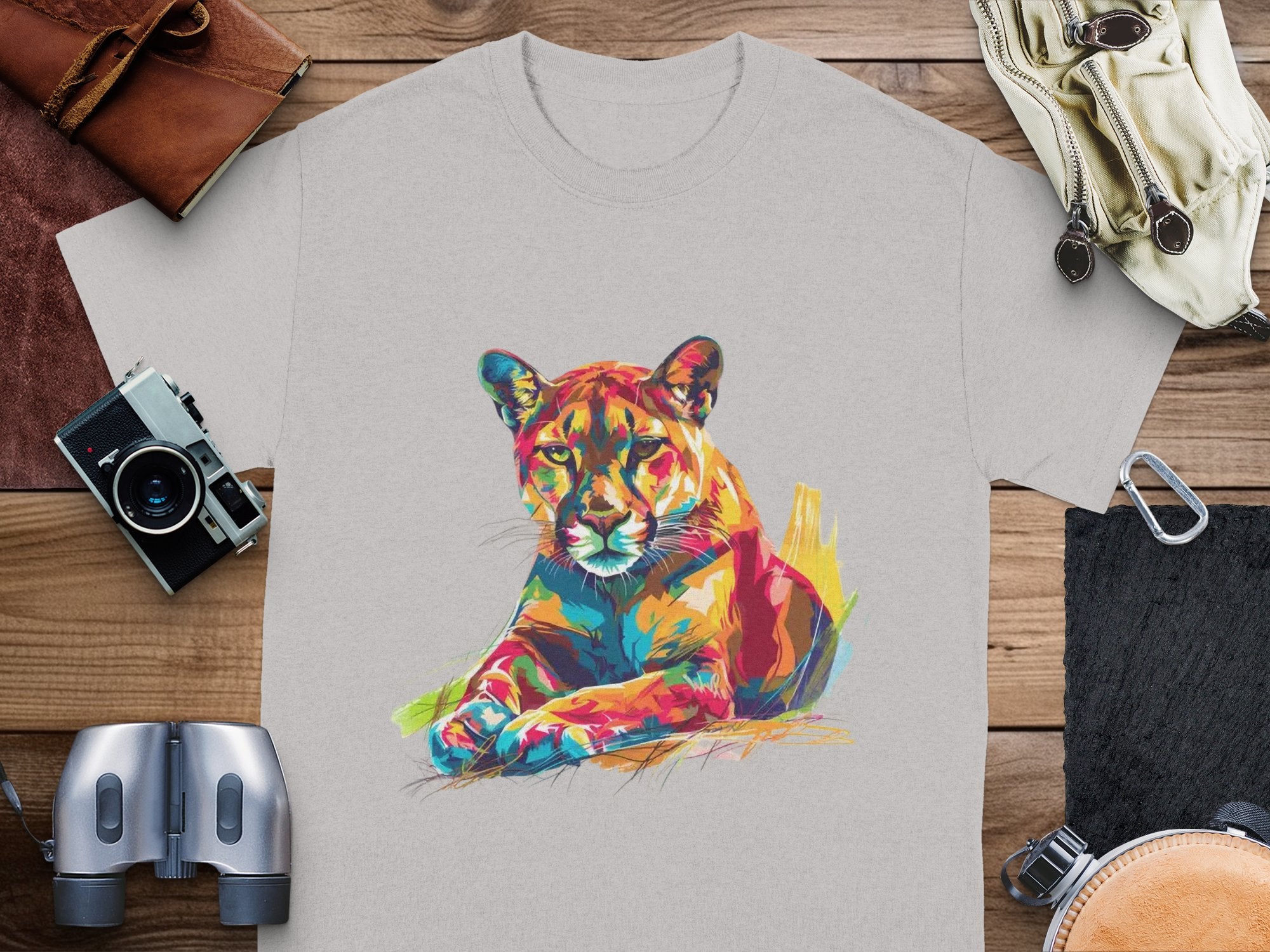 A Garment Graphics light gray graphic print T-shirt showcases a colorful cougar in vibrant artwork. Its displayed on wood, surrounded by a vintage camera, binoculars, leather bag, and gloves.