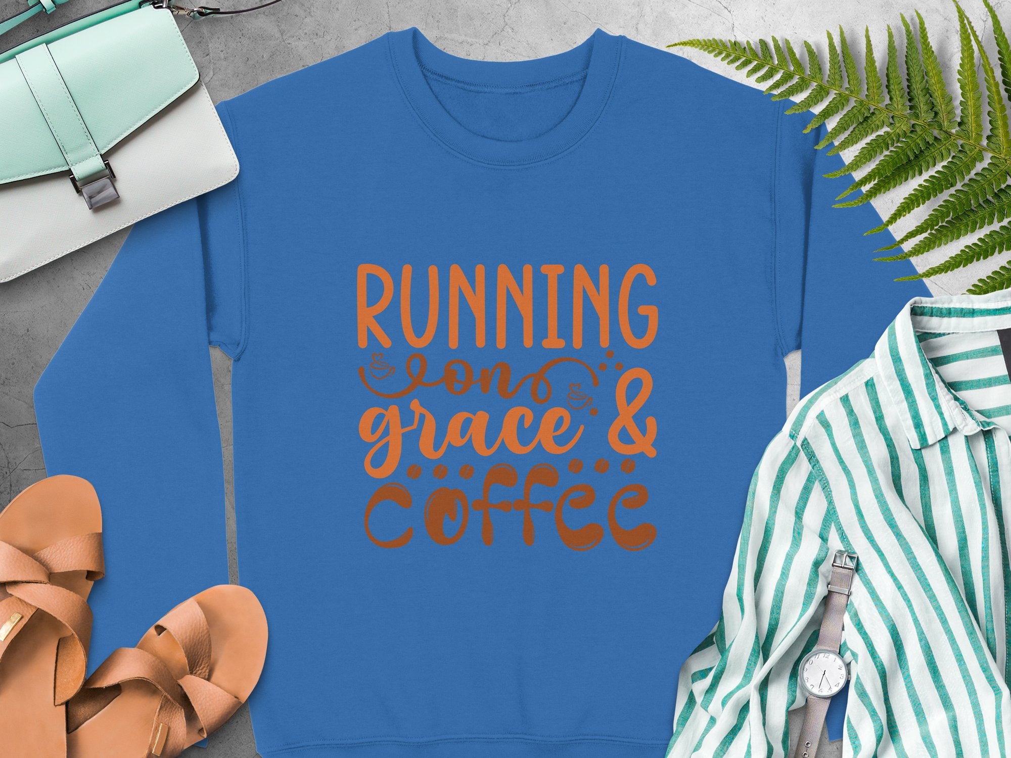 A blue Garment Graphics sweatshirt with Running on Grace & Coffee in orange, surrounded by tan sandals, a light blue purse, a striped blouse, a watch, and fern leaves—all artfully set on a sleek gray surface.