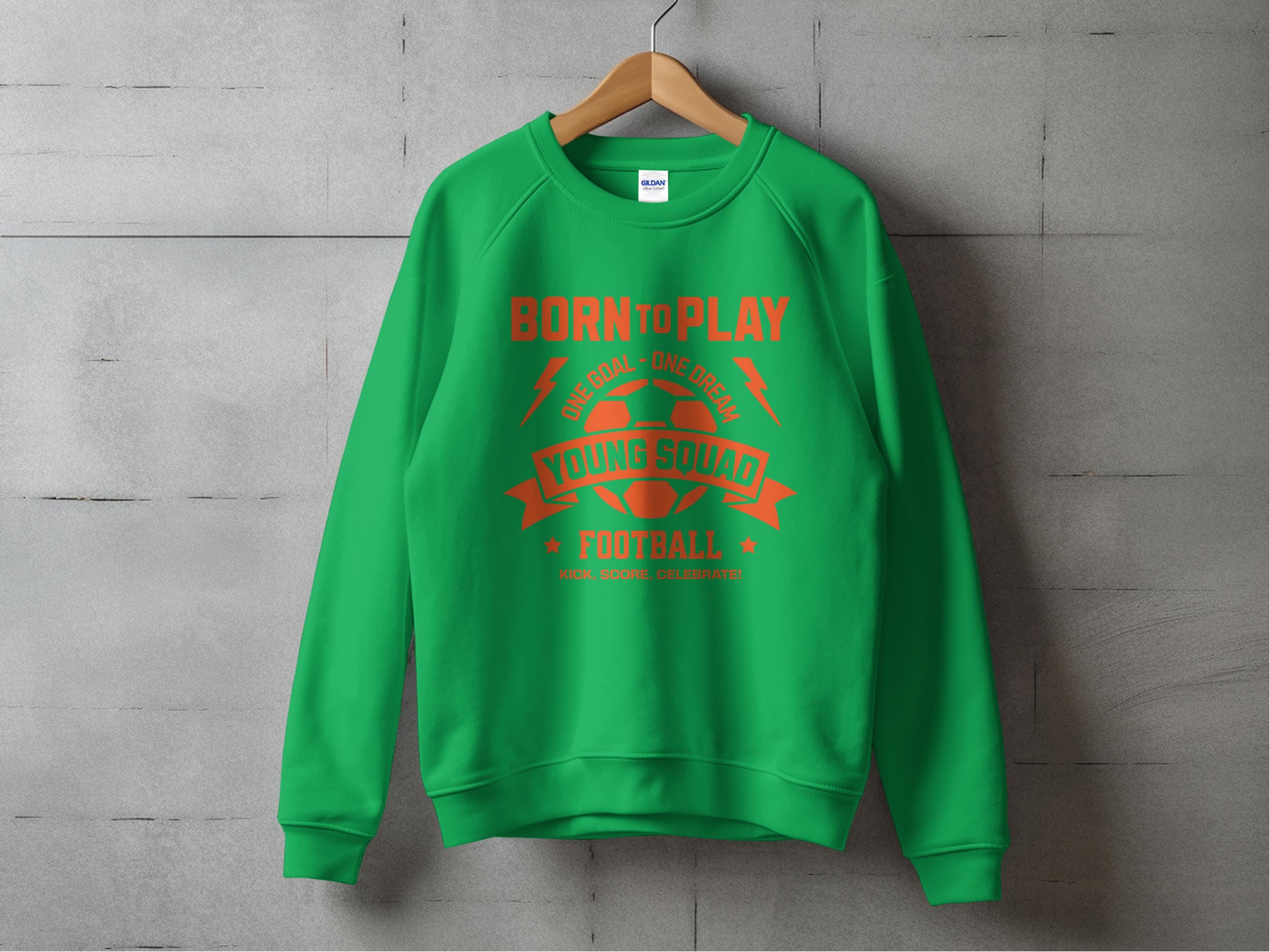 A green football sweatshirt from Garment Graphics hangs on a concrete wall, featuring bold red BORN to PLAY and YOUNG SQUAD FOOTBALL text and graphics. The unisex design is ideal for young aspiring footballers, highlighted by a central football graphic.