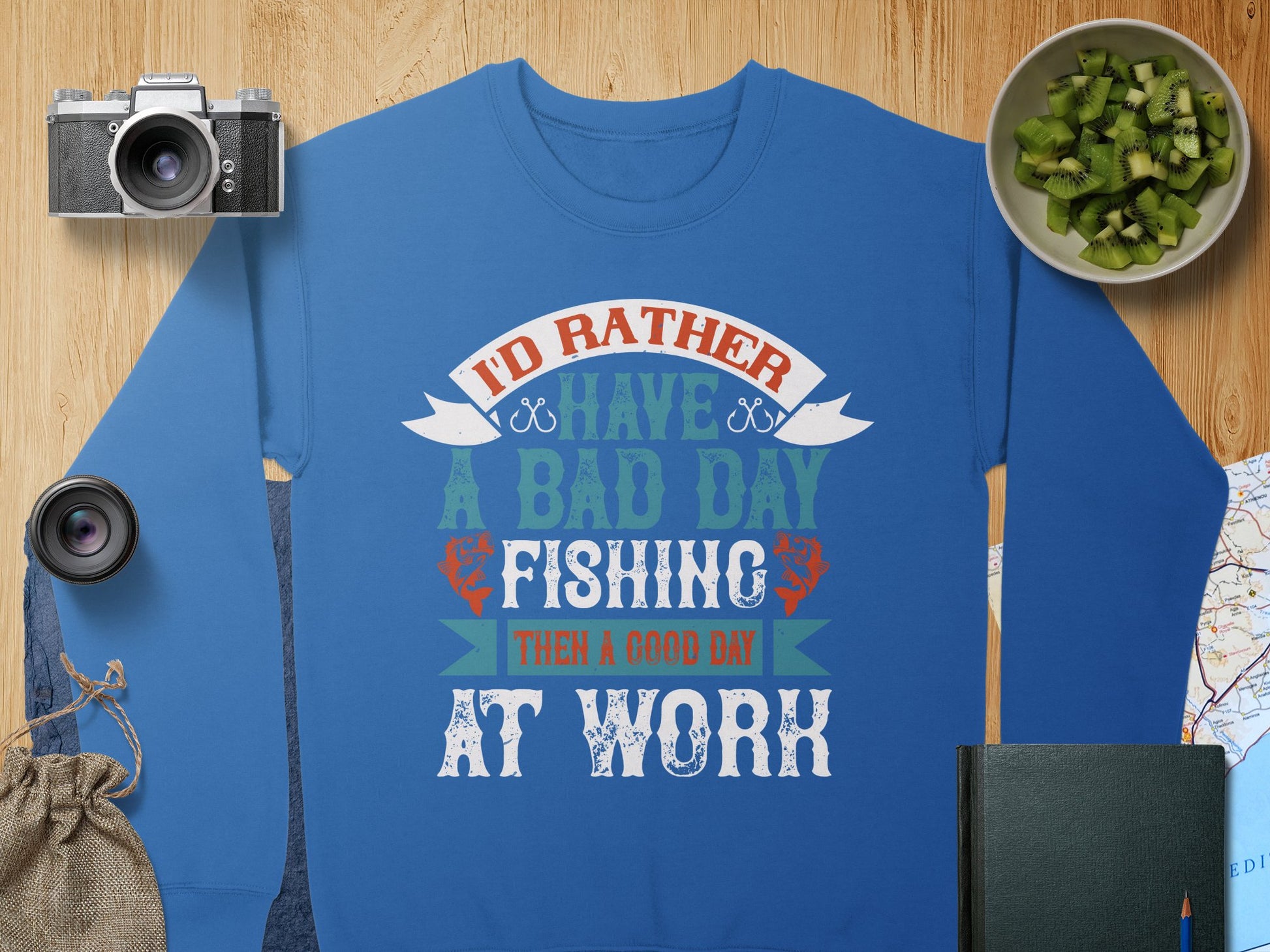 Garment Graphics Unisex Fishing Sweatshirt features the quote Id rather have a bad day fishing than a good day at work in white, orange, and green. Paired with items like a camera, lens, pouch, kiwi bowl, notebook, and map—it’s perfect for expressing your passion for fishing!.