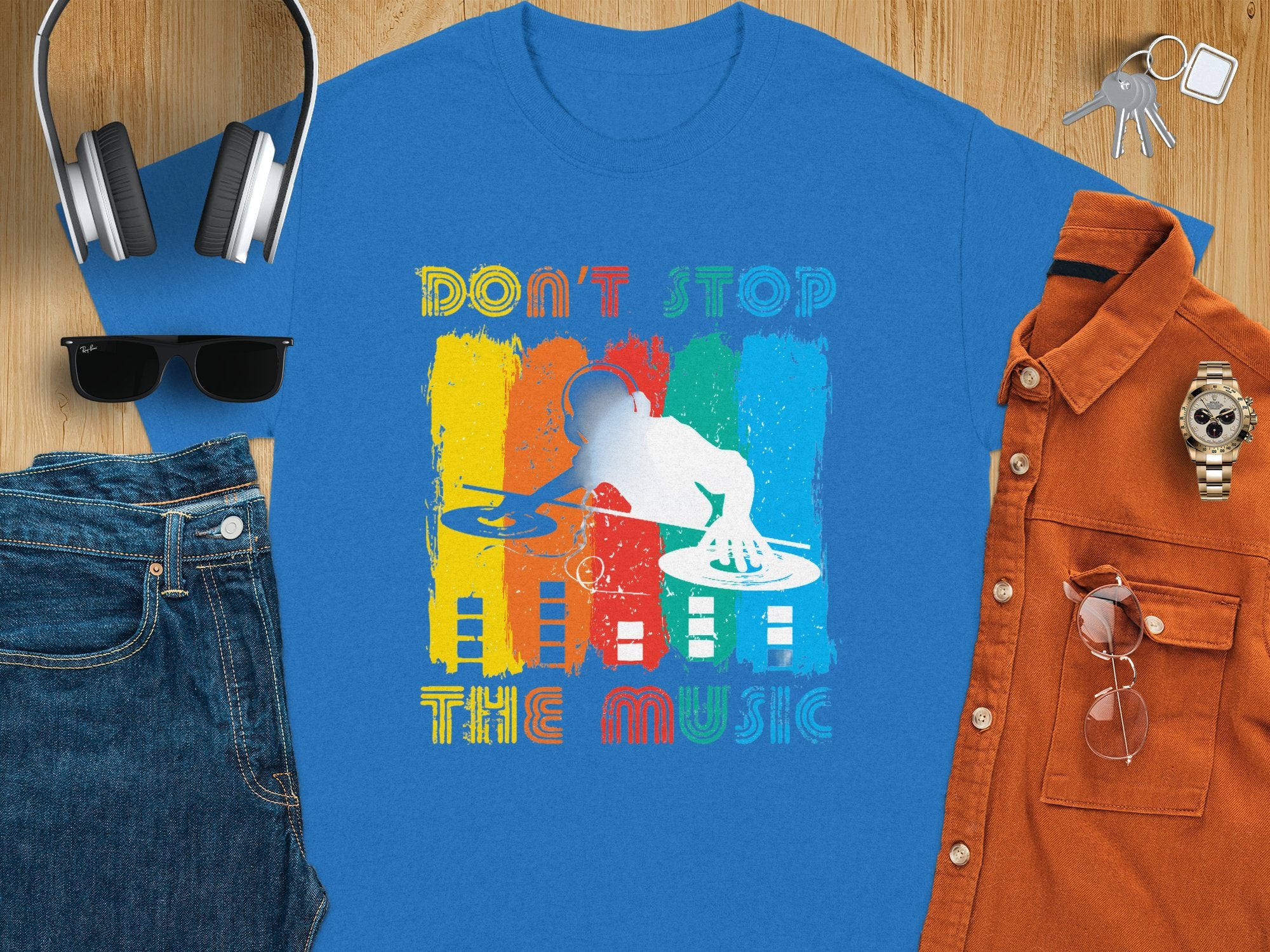 Flat lay of a Garment Graphics blue t-shirt featuring a DJ graphic and Dont Stop the Music text in vibrant colors. Ideal for music lovers and party-goers, its complemented by headphones, a cap, sunglasses, jeans, keys, an orange shirt, and a wristwatch on a wooden surface.