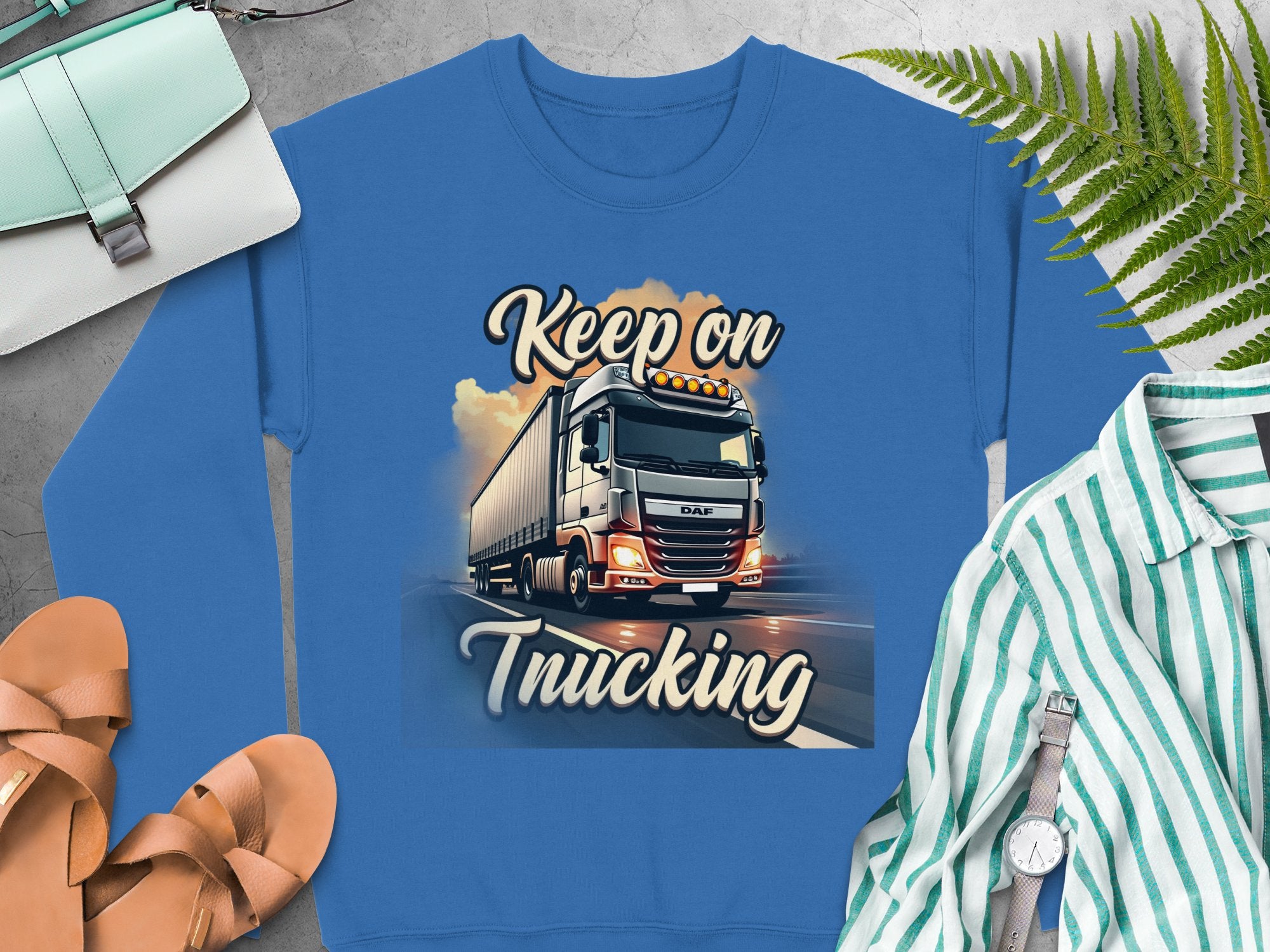 A blue unisex graphic sweatshirt by Garment Graphics, showcasing a large truck with Keep On Trucking boldly printed, is paired with accessories like a handbag, sandals, and a striped shirt amidst ferns on a gray surface.