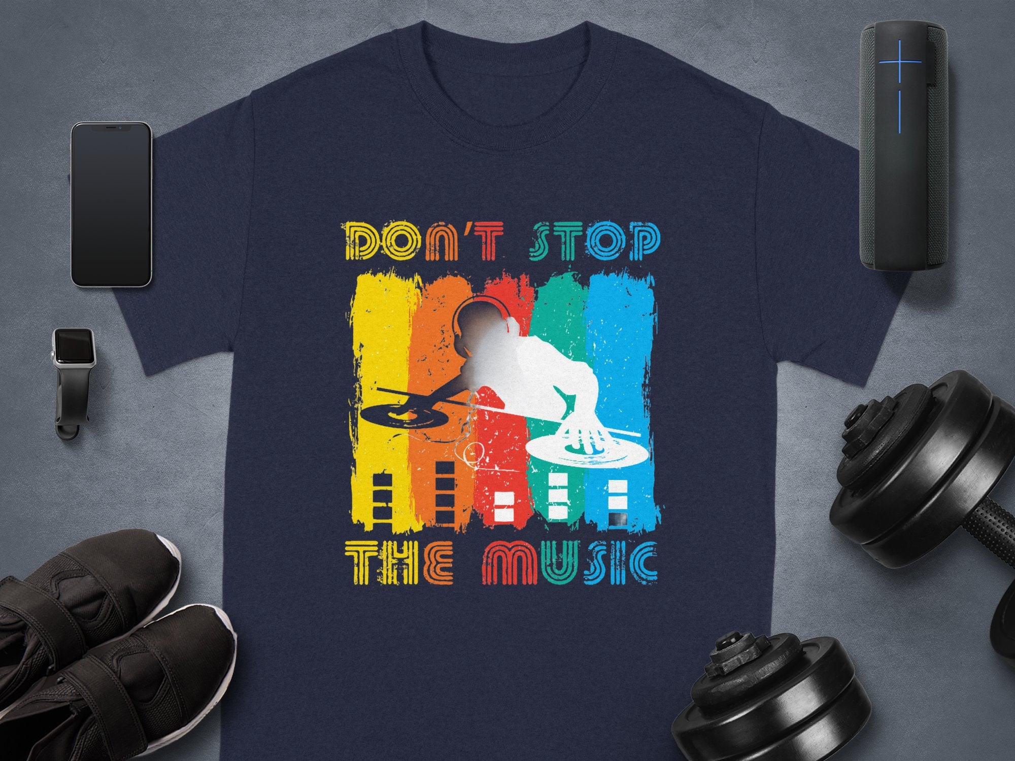 A Garment Graphics T-shirt featuring a DJ graphic and Dont Stop the Music slogan, ideal for music fans, lies on a gray background with a smartphone, smartwatch, shoes, portable speaker, and dumbbells. The design is vibrant with colorful elements.