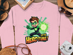 Garment Graphics presents a pink cartoon-themed sweatshirt featuring a superhero boy in a green glowing costume with the slogan Its Hero Time! Unisex sizing ensures a perfect fit. Shown on a wooden surface with sandals, hat, sunglasses, and camera.