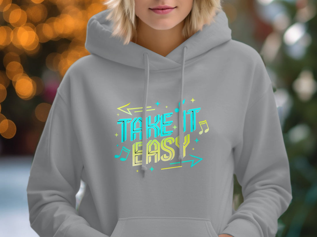 Garment Graphics presents a white hoodie with Take It Easy in vivid blue and yellow. Laid on wood, its accompanied by sunglasses, a sun hat, sandals, a notebook, pen, and camera—embodying relaxed fashion and casual style.