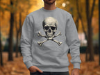 In a vibrant autumn park, someone wears a bold and edgy sweatshirt by Garment Graphics. The light gray sweater features a large skull and crossbones design, contrasting against the rich background of colorful trees and fallen leaves, enhancing its daring vibe.