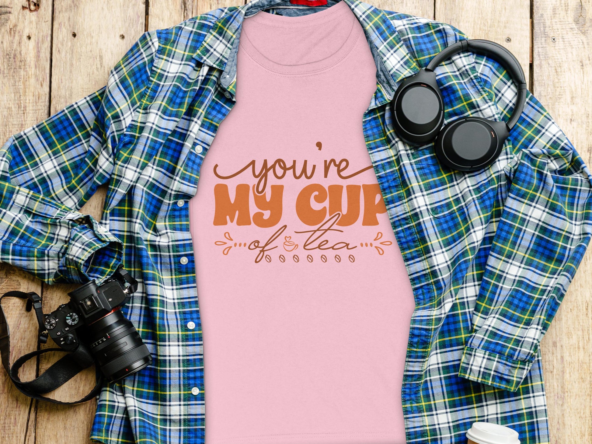 A Garment Graphics 100% cotton graphic T-shirt with Youre My Cup of Tea text in pink lies on a wooden surface, partially covered by a blue and green plaid cloth. Its accompanied by charming finds like a camera, headphones, and a coffee cup.