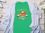 The Garment Graphics T-shirt showcases a cartoon pie with whipped cream, Hot & Spicy text above, and You Wanna a Piece of Me? below. Ideal for family sizing, it’s displayed on wood next to a light gray cardigan.