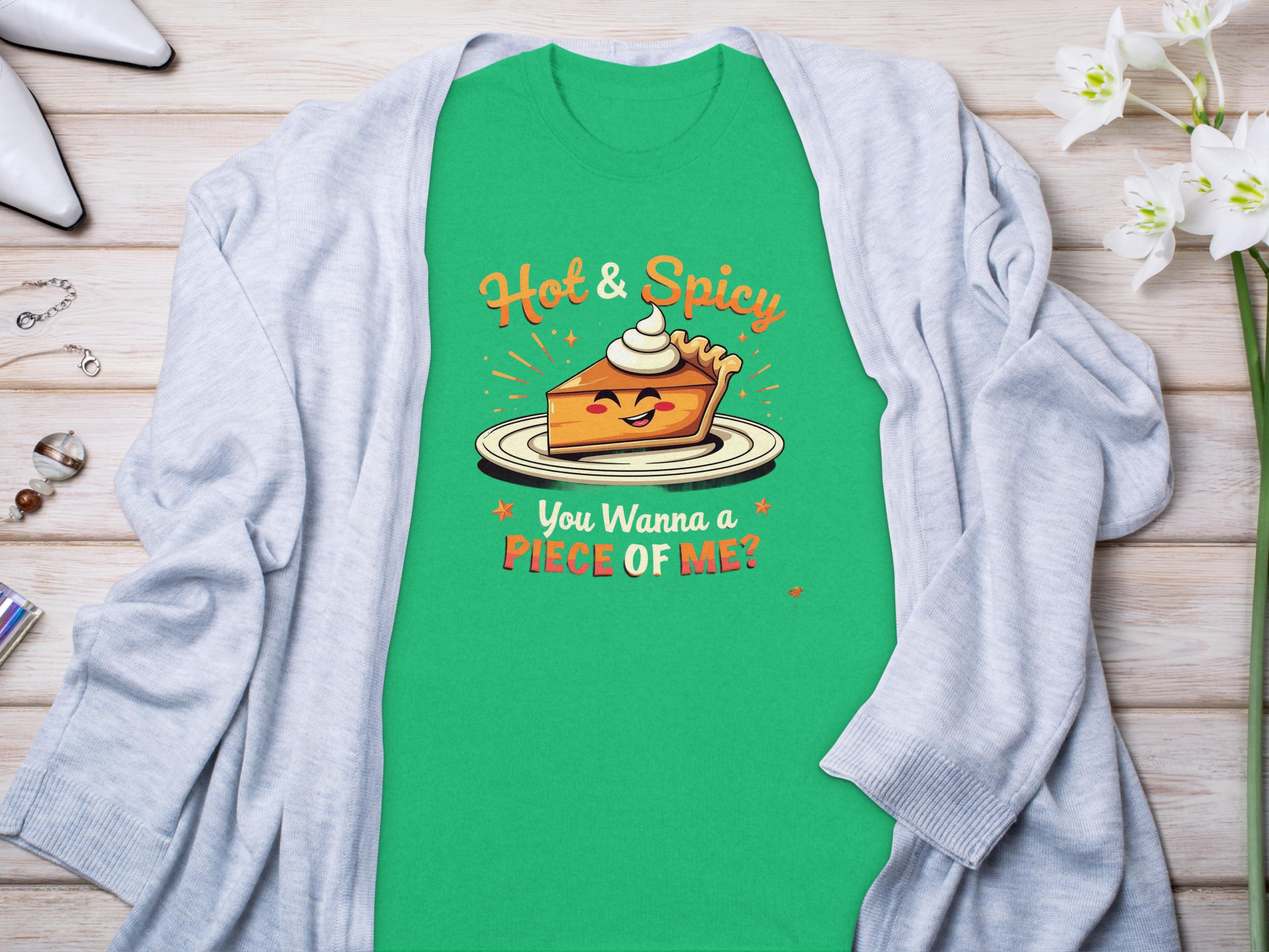 The Garment Graphics T-shirt showcases a cartoon pie with whipped cream, Hot & Spicy text above, and You Wanna a Piece of Me? below. Ideal for family sizing, it’s displayed on wood next to a light gray cardigan.
