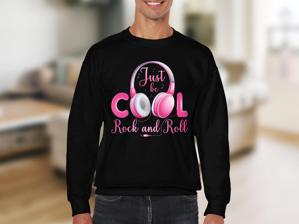 A person wearing a black Garment Graphics sweatshirt with Just be Cool Rock and Roll text and a pink and white headphones design creates a fashionable statement piece that captivates music lovers in a blurred interior setting.