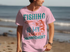 A fishing enthusiast at the beach wears a Garment Graphics pink T-shirt with Fishing is My Anger Management, alongside a fish and fishing lure design.