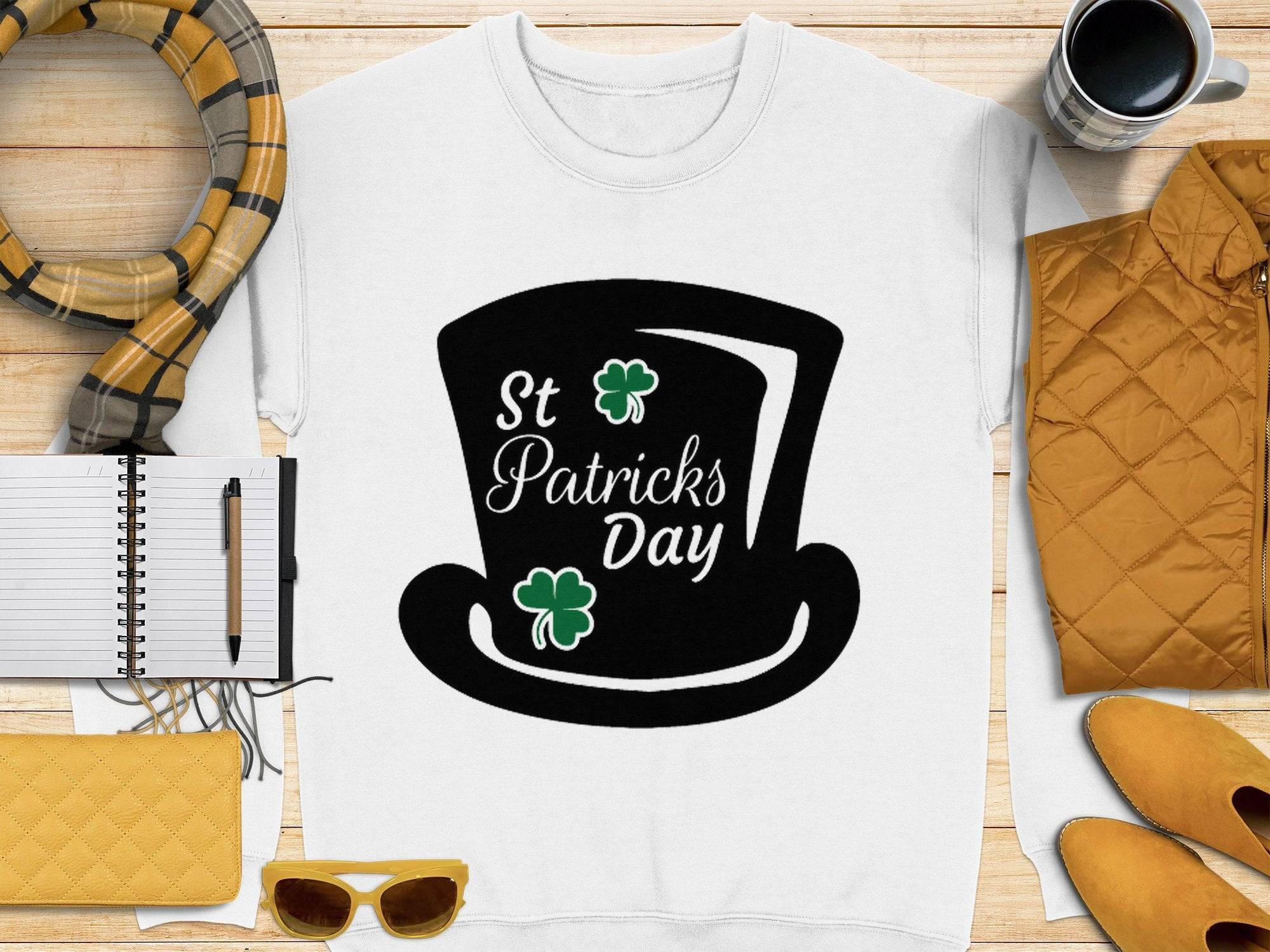 A flat lay image showcases Garment Graphics stylish sweatshirt featuring a black top hat graphic with St Patricks Day and green shamrocks, paired with yellow sunglasses, a scarf, a pen on a notebook, a yellow bag, a jacket, and a coffee cup to capture the festive spirit.