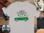 Garment Graphics offers a gray T-shirt with a green pickup truck loaded with shamrocks and Happy St. Patricks Day text, surrounded by a camera, binoculars, wallet, and more on a wooden surface to celebrate Irish pride.