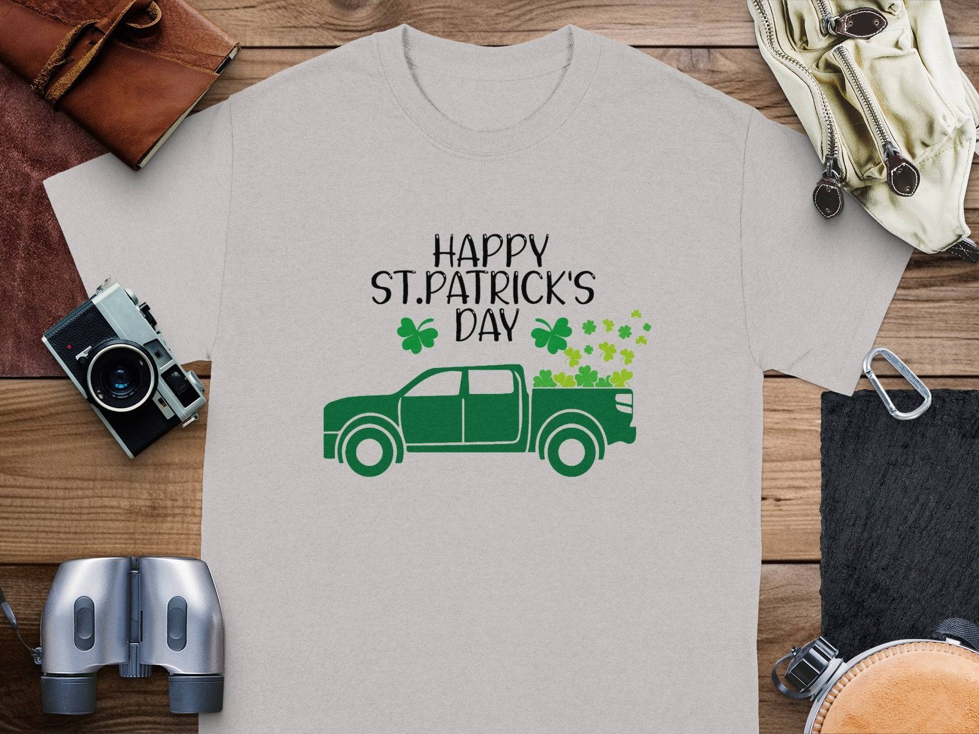 Garment Graphics offers a gray T-shirt with a green pickup truck loaded with shamrocks and Happy St. Patricks Day text, surrounded by a camera, binoculars, wallet, and more on a wooden surface to celebrate Irish pride.