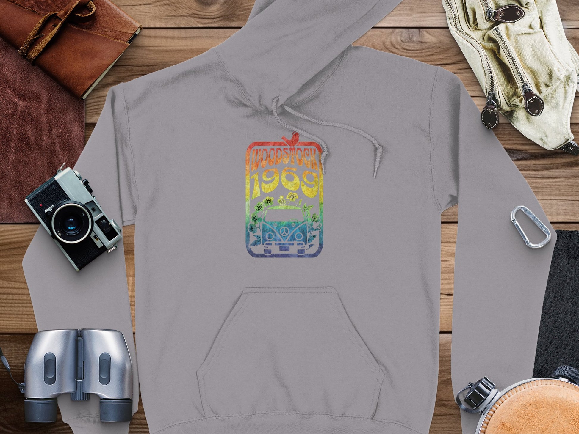 A Garment Graphics gray hoodie with Woodstock 1969 in rainbow colors above a retro bus lies on wood, surrounded by a camera, binoculars, gloves, and a carabiner.