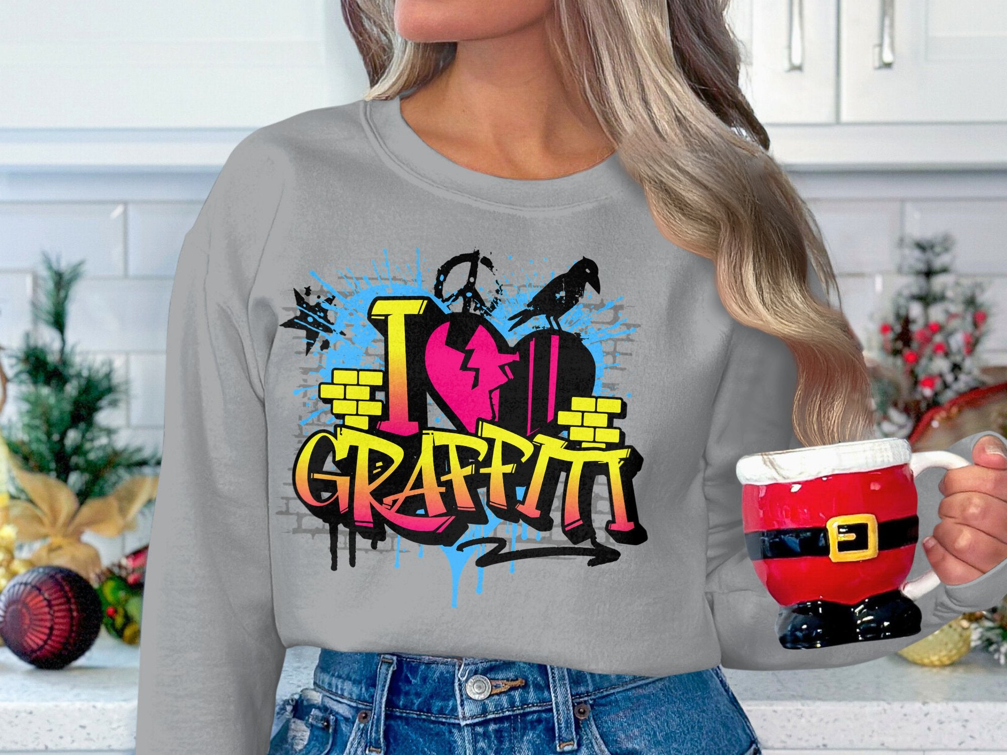 In a festive kitchen, someone holds a Santa-themed mug while wearing Garment Graphics grey sweatshirt, featuring the vibrant I Love Graffiti design with a colorful heart and paint splatters that showcase urban style.