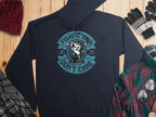 A navy graphic hoodie from Garment Graphics with a circular fish design and Fishing Hair Dont Care text is laid flat on wood. Ideal for anglers, its paired with a maroon beanie, plaid shirt, black and white gloves, and a gray plaid scarf.