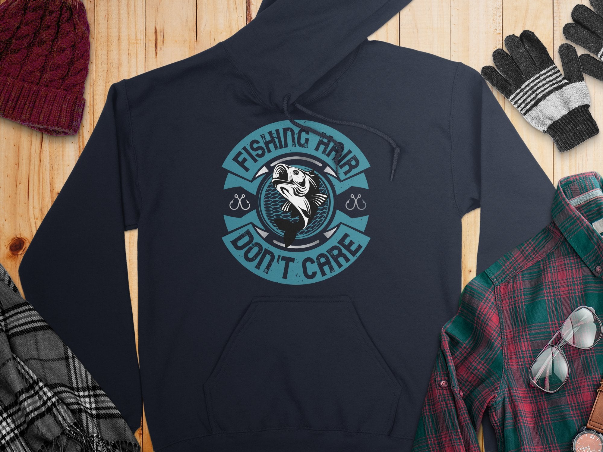A navy graphic hoodie from Garment Graphics with a circular fish design and Fishing Hair Dont Care text is laid flat on wood. Ideal for anglers, its paired with a maroon beanie, plaid shirt, black and white gloves, and a gray plaid scarf.