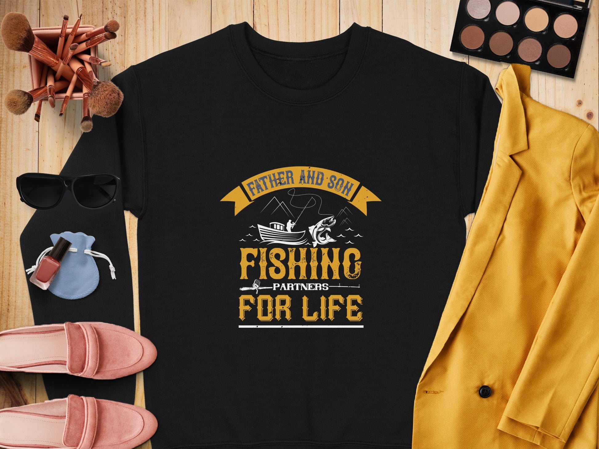 Flat lay showcasing a black Garment Graphics sweatshirt with Father and Son Fishing Partners for Life text, surrounded by a yellow jacket, pink loafers, makeup brushes, eyeshadow palette, sunglasses, watch, nail polish, and blue pouch on wood.